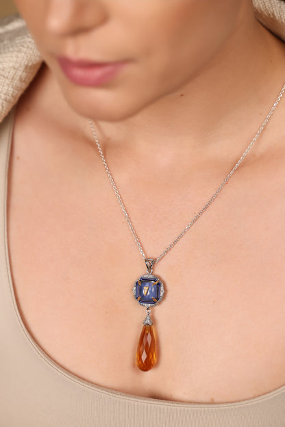 A close-up of a model wearing a silver chain necklace with a teardrop-shaped amber gemstone pendant and a square blue gemstone accent.