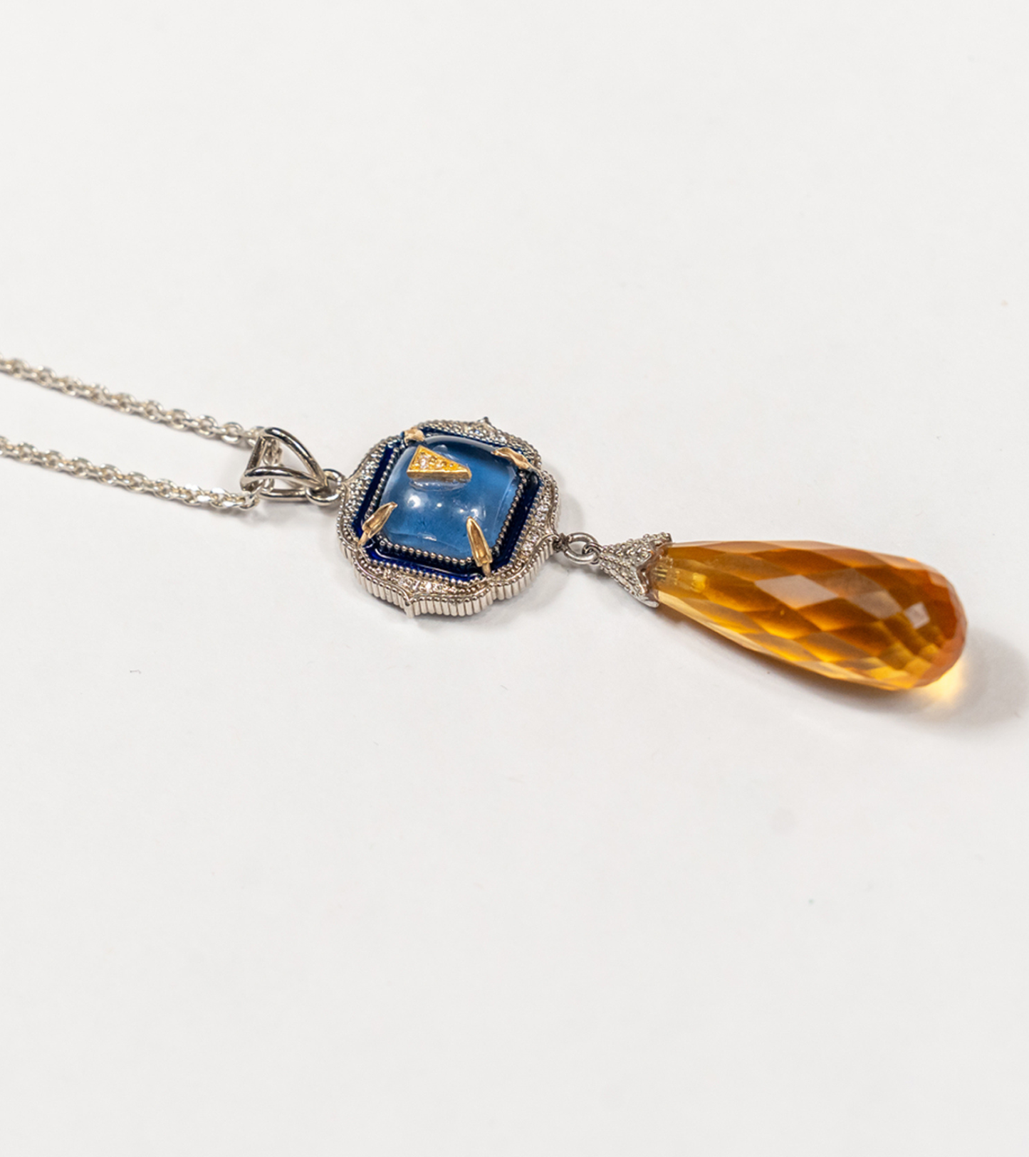 A silver pendant necklace featuring a square blue enamel medallion with gold detailing, suspended from a silver chain and connected to a faceted amber-orange teardrop crystal drop, shown on a plain white background.