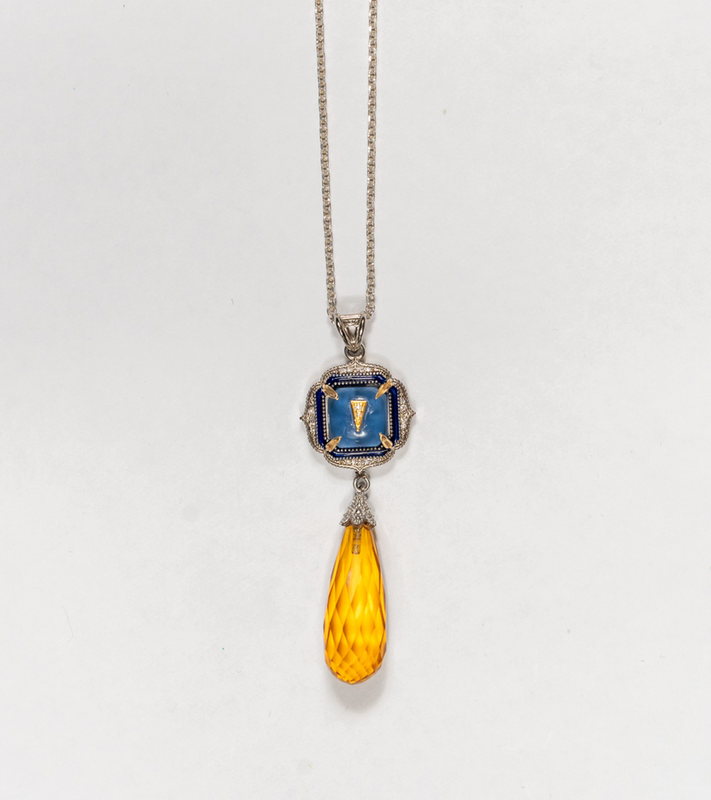 A silver pendant necklace featuring a square blue enamel medallion with a gold Roman numeral 'I' and a faceted amber-yellow teardrop crystal drop, shown against a plain white background.