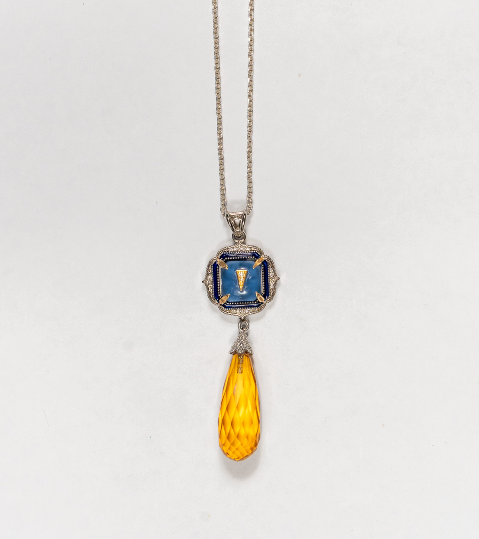 A silver pendant necklace featuring a square blue enamel medallion with a gold Roman numeral 'I' and a faceted amber-yellow teardrop crystal drop, shown against a plain white background.
