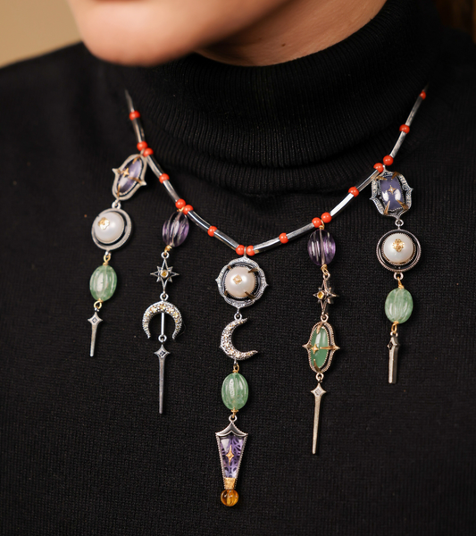 Close-up of the Amdavad Necklace's dramatic pendants — crescent moon, violet enamel dagger, and star motifs — against the coral bead collar